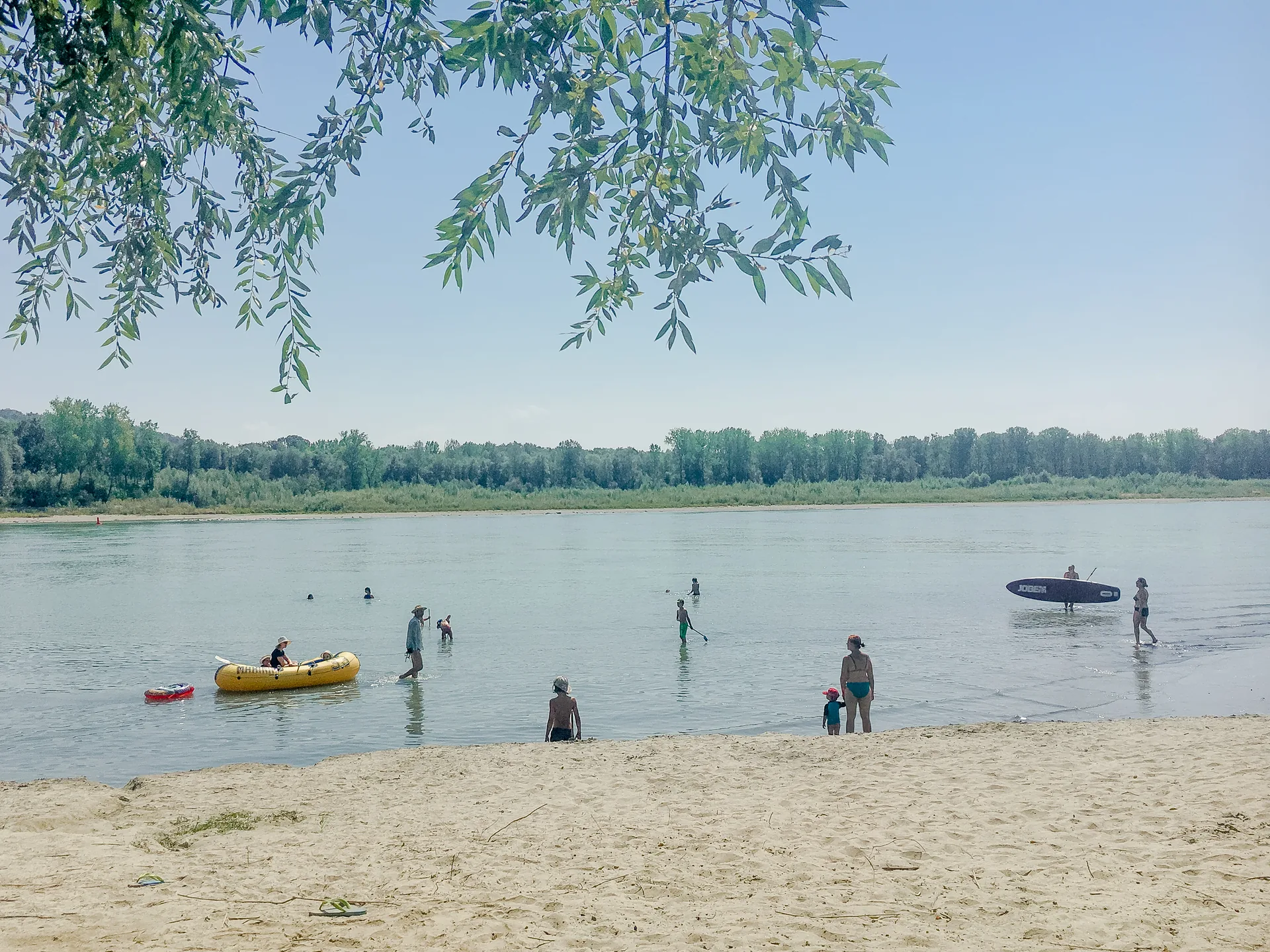 Flussstrand mit Badegästen und Booten unter Weidenzweigen