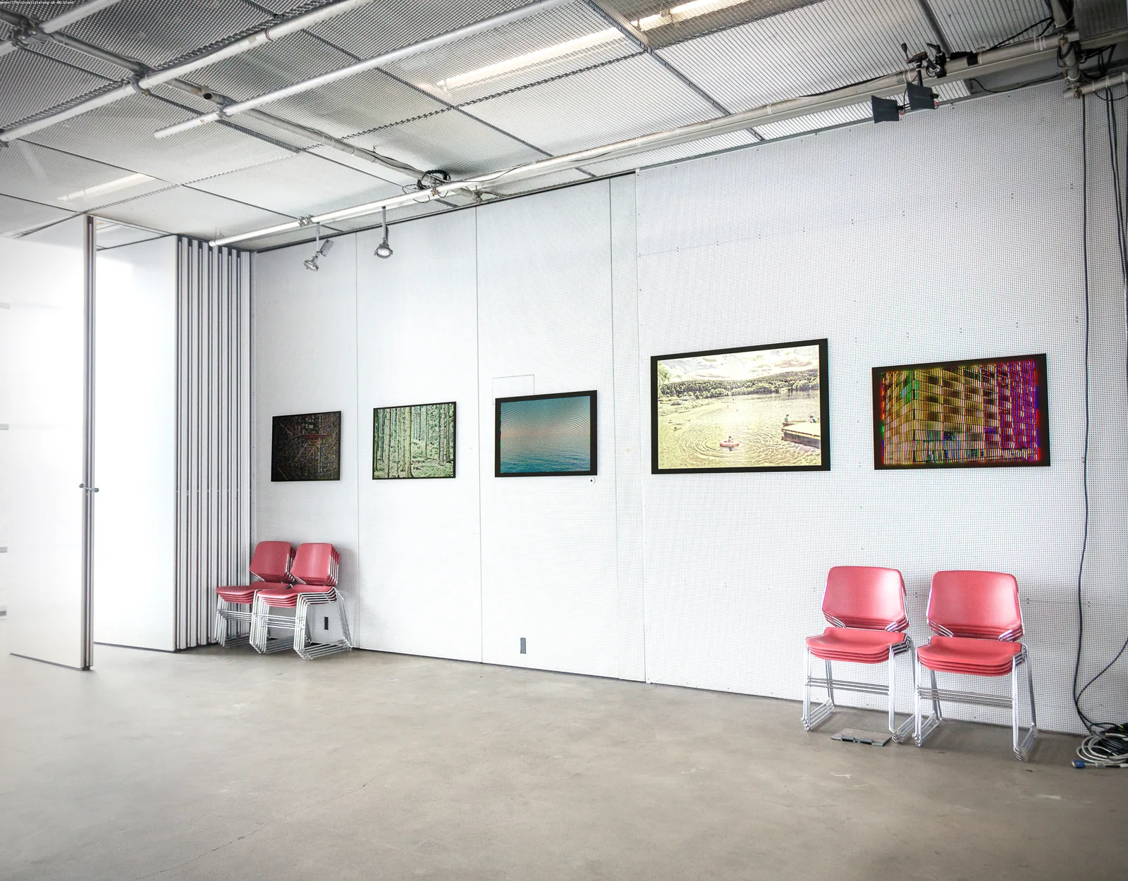 Ausstellungsraum mit fünf gerahmten Bildern an weißer Wand, davor rote Stühle