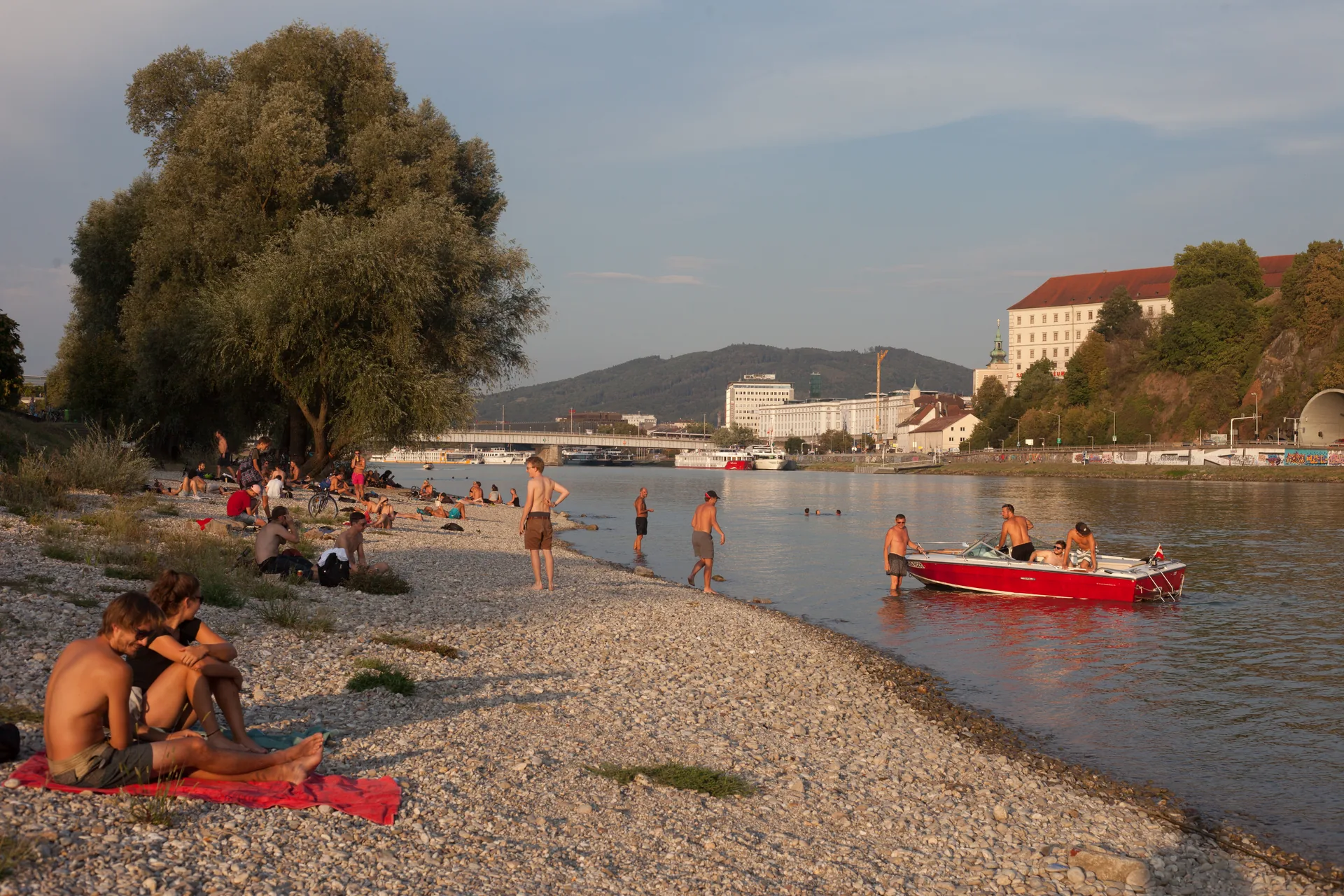 Kiesstrand an der Donau bei Linz mit Badegästen und rotem Motorboot am Abend