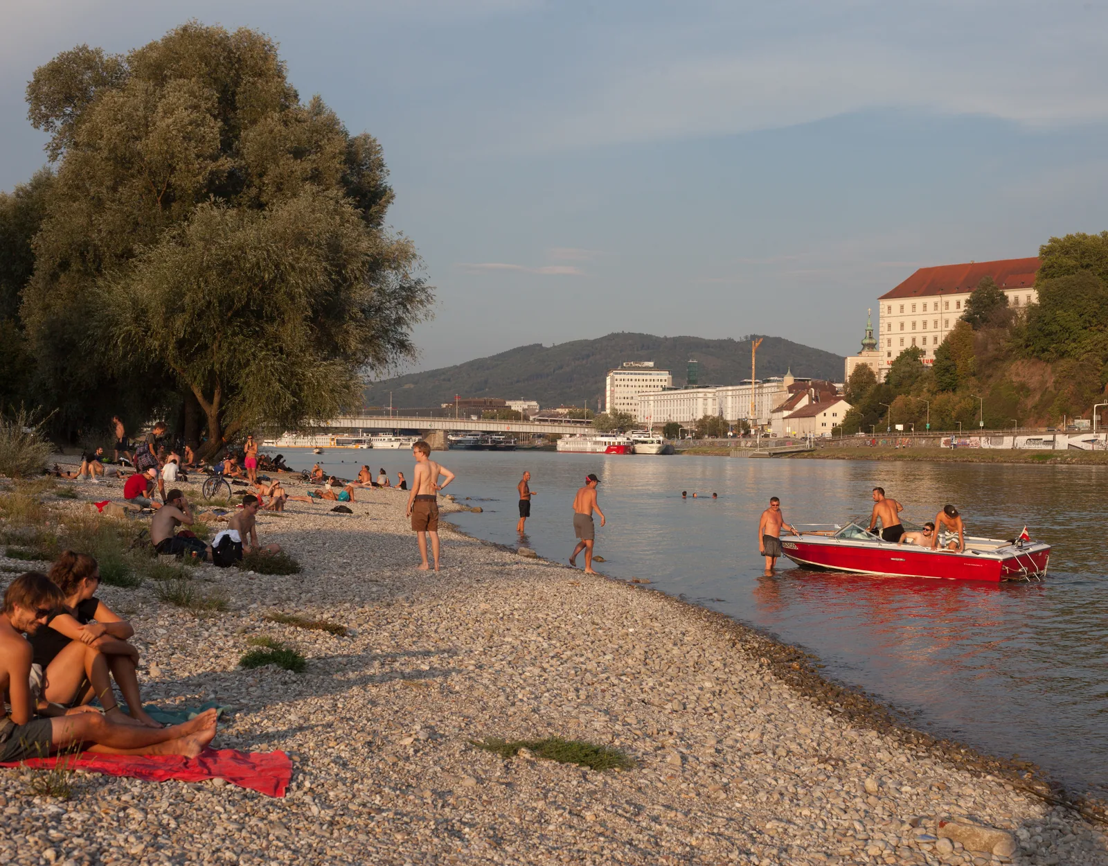 Sommerabend an der Donau: Badegäste am Kiesstrand, rotes Boot, Linz im Hintergrund